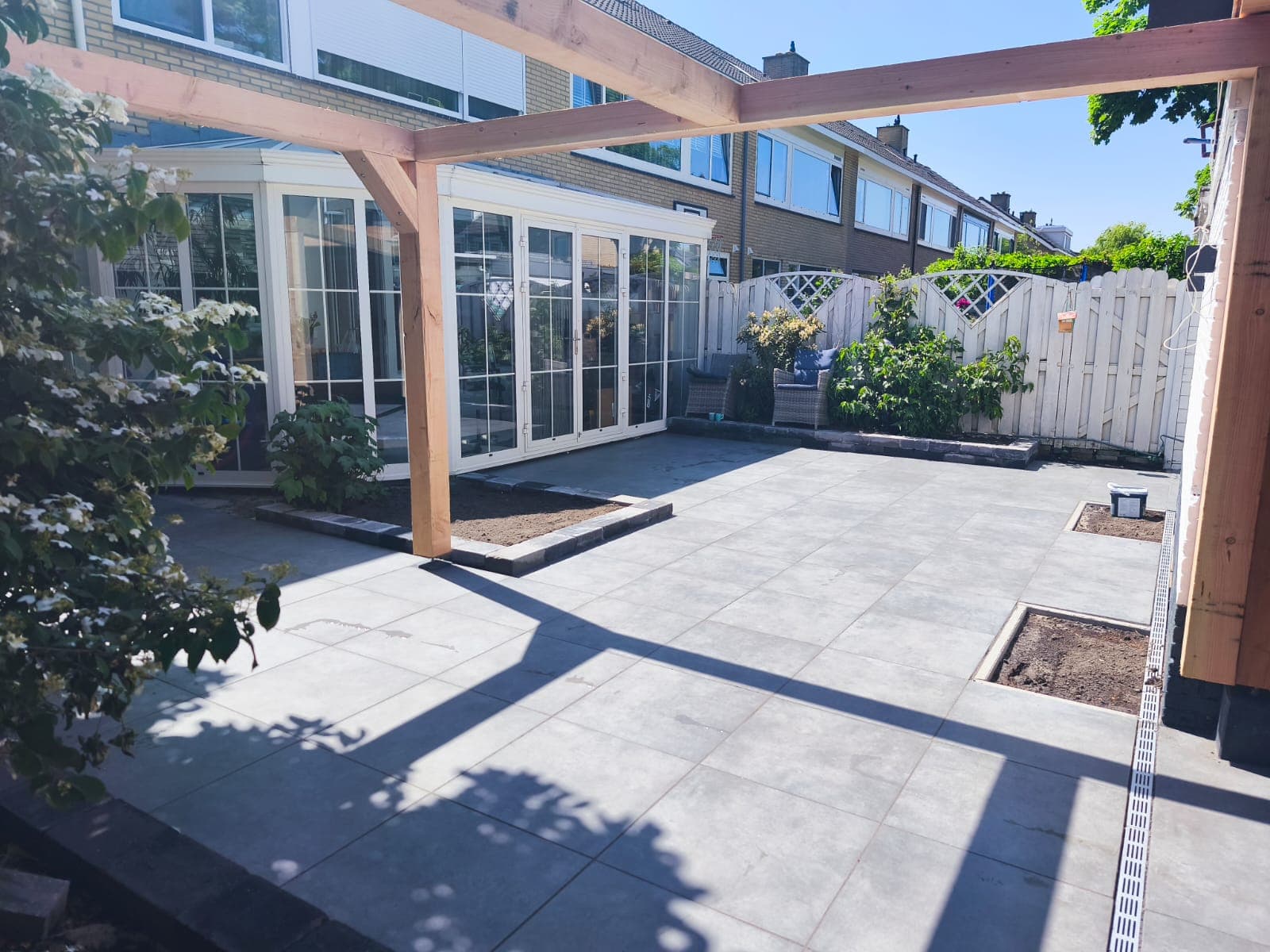 Grey porcelain patio under a new Douglas fir pergola, looking toward a white-framed conservatory